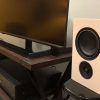 Beige PSB IQ2 wireless speakers sit on stands near a TV