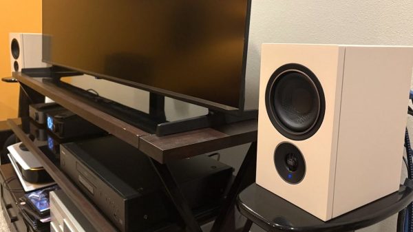 Beige PSB IQ2 wireless speakers sit on stands near a TV